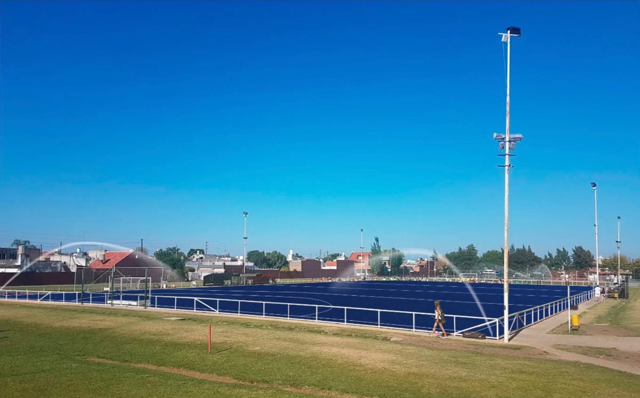 Cancha de hockey sintético de agua azul con los aspersores funcionando, tomada como referencia.